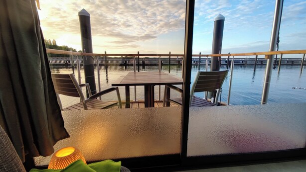A view from a room with a large window overlooking a wooden deck with a table and chairs, a railing, and a body of water with a dock in the background.