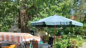 An outdoor patio with a table and chairs under a green umbrella, surrounded by potted plants and a gravel path.