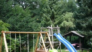 A wooden play structure with swings and slides in a grassy area surrounded by trees.