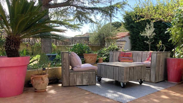 Un patio al aire libre con un sofá y una mesa de madera, rodeado de macetas con plantas y una palmera.