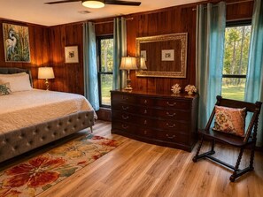 Un dormitorio con cama, cómoda, silla y ventanas amplias.