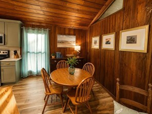 A wooden-paneled room with a dining table, chairs, a lamp, and framed pictures on the wall.