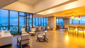 A modern hotel lobby with large windows offering city views, a bar area with seating, and a spacious seating arrangement with sofas and chairs.