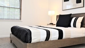 A neatly made bed with a black and white striped bedspread, a bedside table with a lamp, and a window with blinds. - Spacious Condo in Southfield, Michigan (Southfield)
