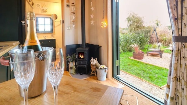 A cozy living room with a fireplace, a large window, and a bottle of champagne on the table. - Shepherd's Hut- Hot Tub- Wellness Spa- Wood Burner (Nottingham)