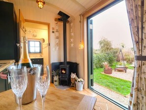 A cozy living room with a fireplace, a large window, and a bottle of champagne on the table. - Shepherd's Hut- Hot Tub- Wellness Spa- Wood Burner (Nottingham)