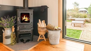A modern interior with a wood-burning stove, a potted plant, a candle holder, and a view of the outdoors. - Shepherd's Hut- Hot Tub- Wellness Spa- Wood Burner (Nottingham)