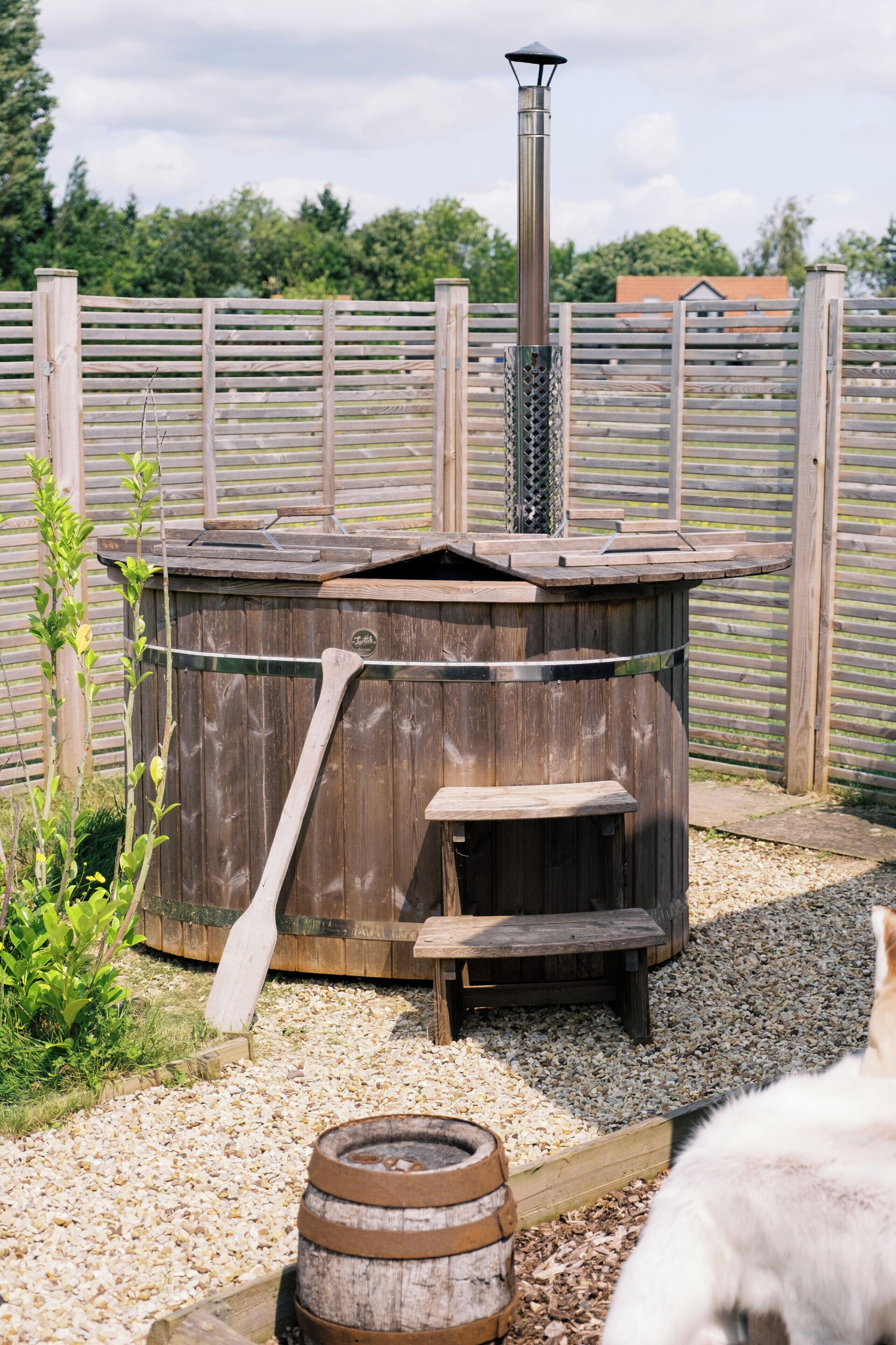 A wooden hot tub with a chimney, surrounded by a wooden fence and gravel.