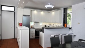 A compact kitchen with white cabinets, a central island with bar stools, a dining table, and a window with blinds. - A private,family-friendly estate with pool, garden,and all comforts—near the sea (Faros Avlidas)