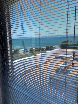 A view through window blinds to a patio area with outdoor furniture and a distant view of the sea.