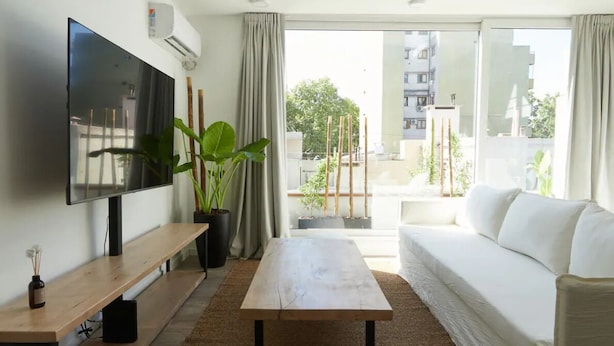 Una sala de estar moderna con un televisor de pantalla plana, un sofá blanco, una mesa de centro de madera y plantas en macetas.