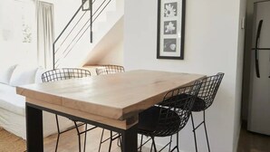 Un comedor con una mesa de madera y sillas negras de alambre, una escalera con barandilla metálica y un refrigerador al fondo.