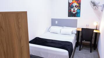 Une chambre à coucher avec un lit, un bureau avec une chaise, un ventilateur fixé au mur et un tableau.
