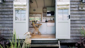 Una cabaña acogedora con fachada de madera, una pequeña cocina con armarios blancos y una zona de comedor con una mesa y sillas.