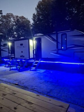 Adjacent RVs sit on a wooden deck surrounded by trees.