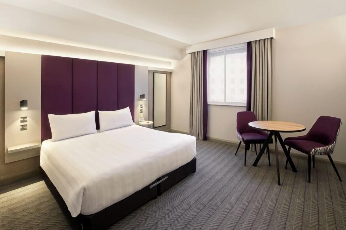 A hotel room with a large bed, a desk with two chairs, a window with curtains, and a wall-mounted mirror.