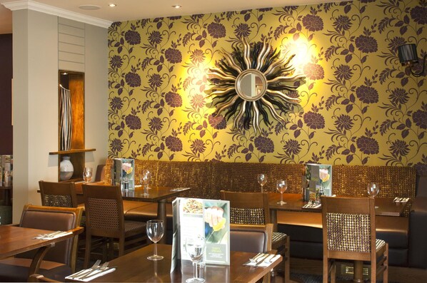 A restaurant interior with wooden tables, chairs, a floral wallpaper design, and a sun-shaped mirror on the wall.