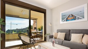 En moderne stue med en grå sofa, en sort fletstol, et hvidt sofabord med vinglas og et stort vindue med udsigt til en balkon og udendørs spiseplads.