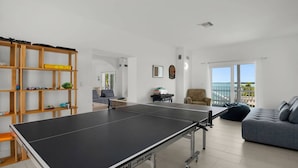 Een moderne woonkamer met een tafeltennistafel, een boekenkast, een bank en uitzicht op zee door grote ramen.