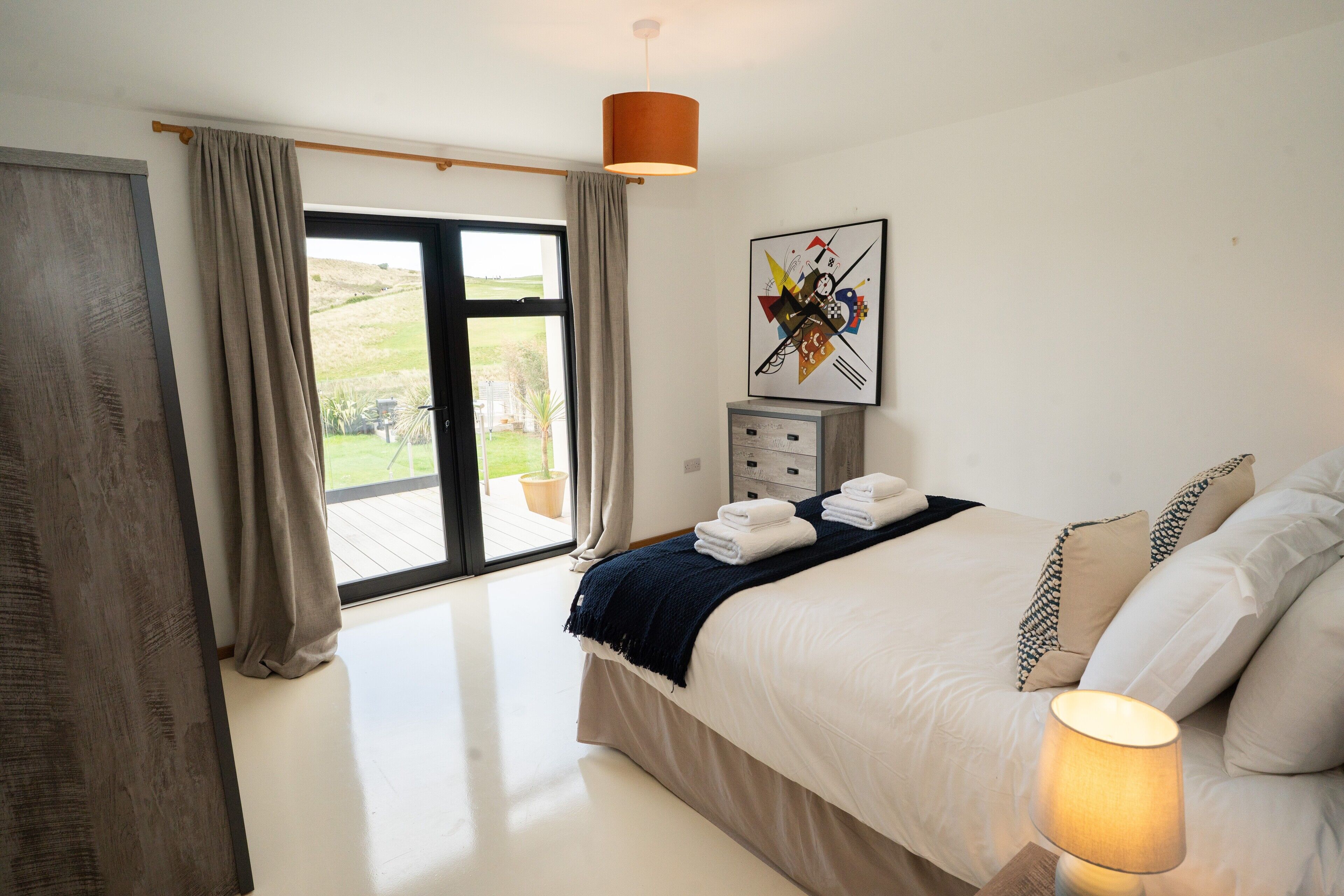 A bedroom with a bed, a bedside table, a lamp, a window with curtains, and a painting on the wall.