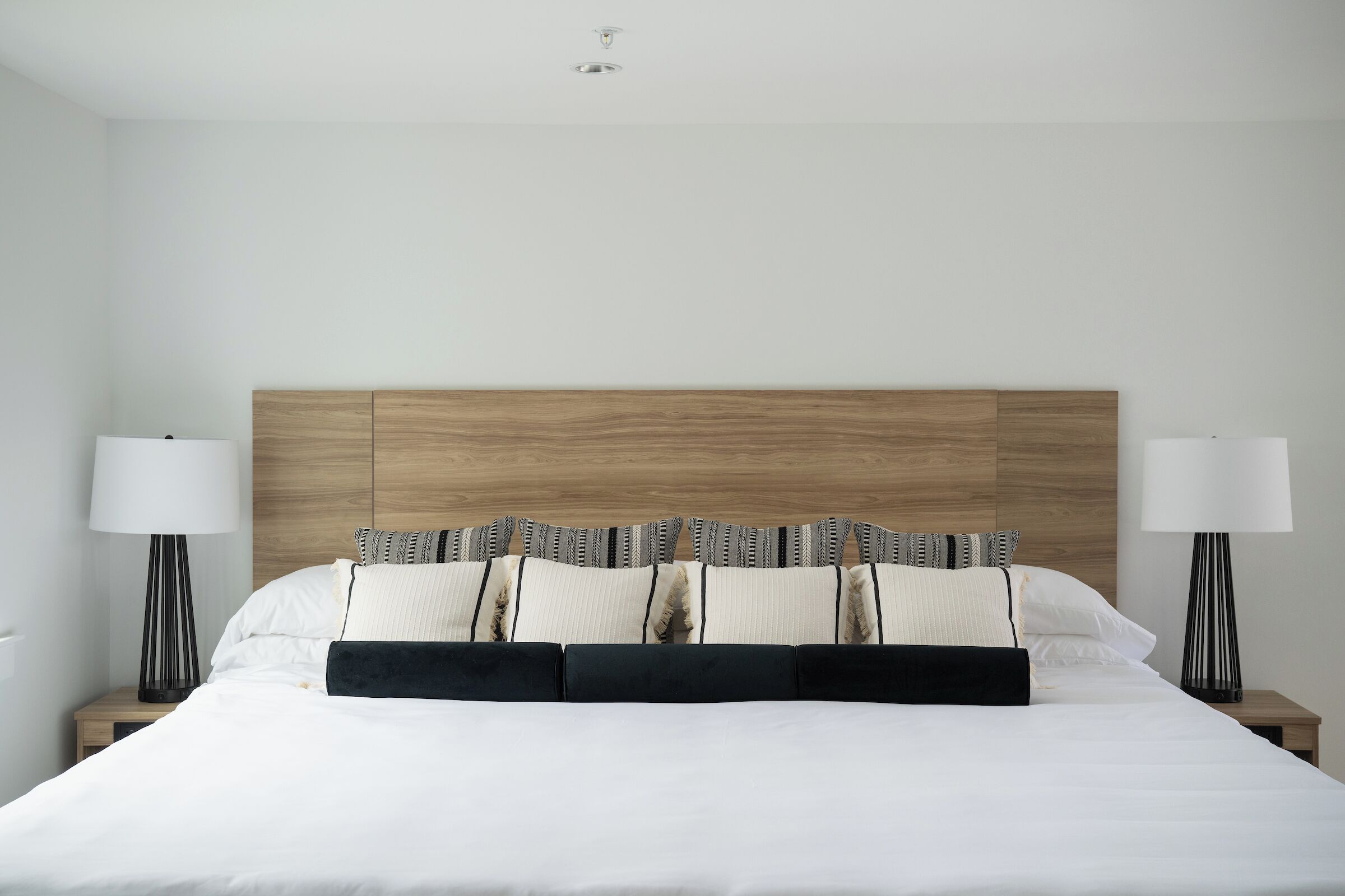A neatly made bed with a wooden headboard, white bedding, and a series of patterned pillows. There are two bedside tables with lamps on either side of the bed.