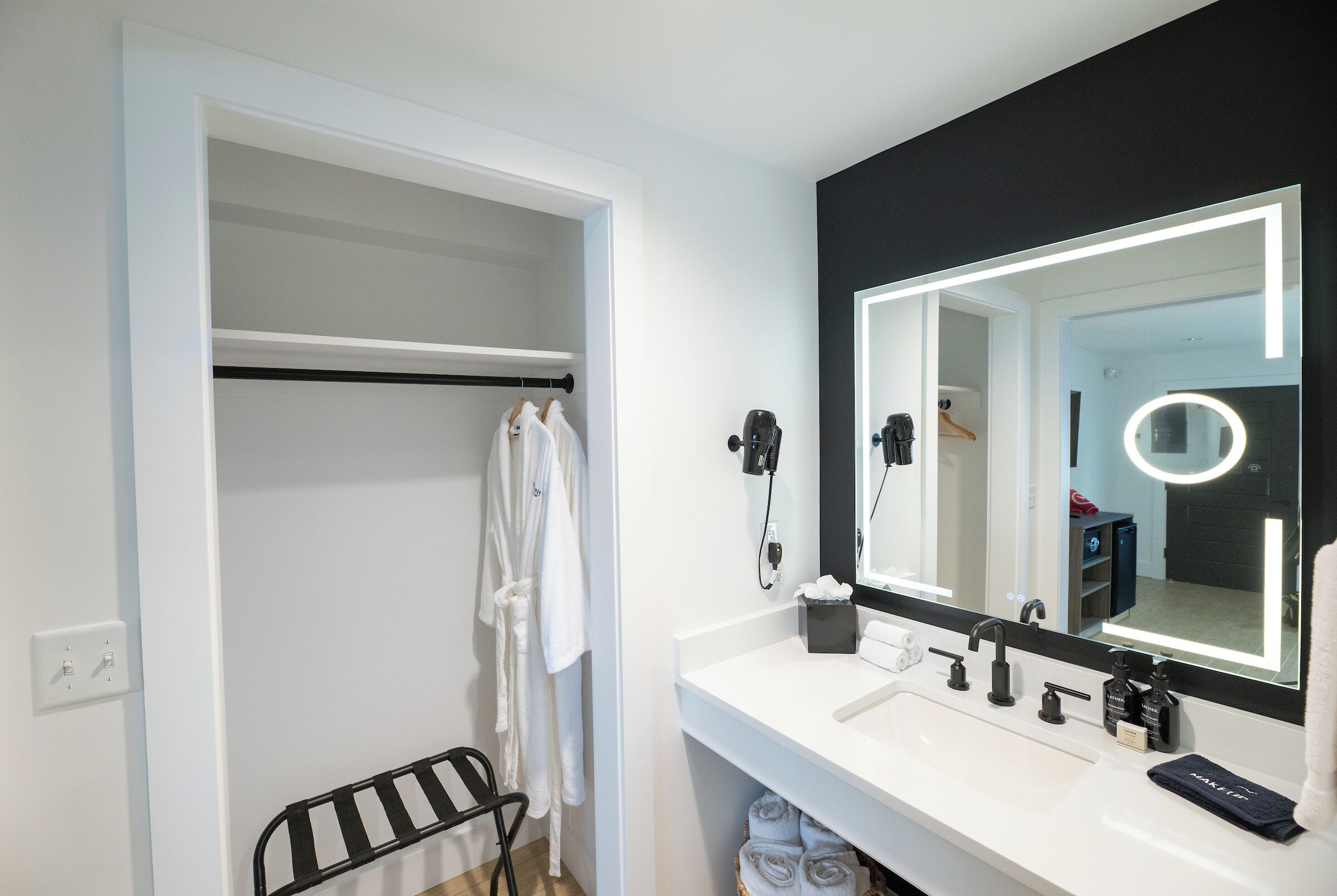 Un baño moderno con un tocador blanco, un espejo grande con iluminación integrada, un perchero negro y una silla negra.