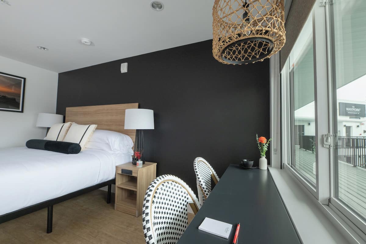 A hotel room with a bed, bedside table, lamp, and a view of the outside.