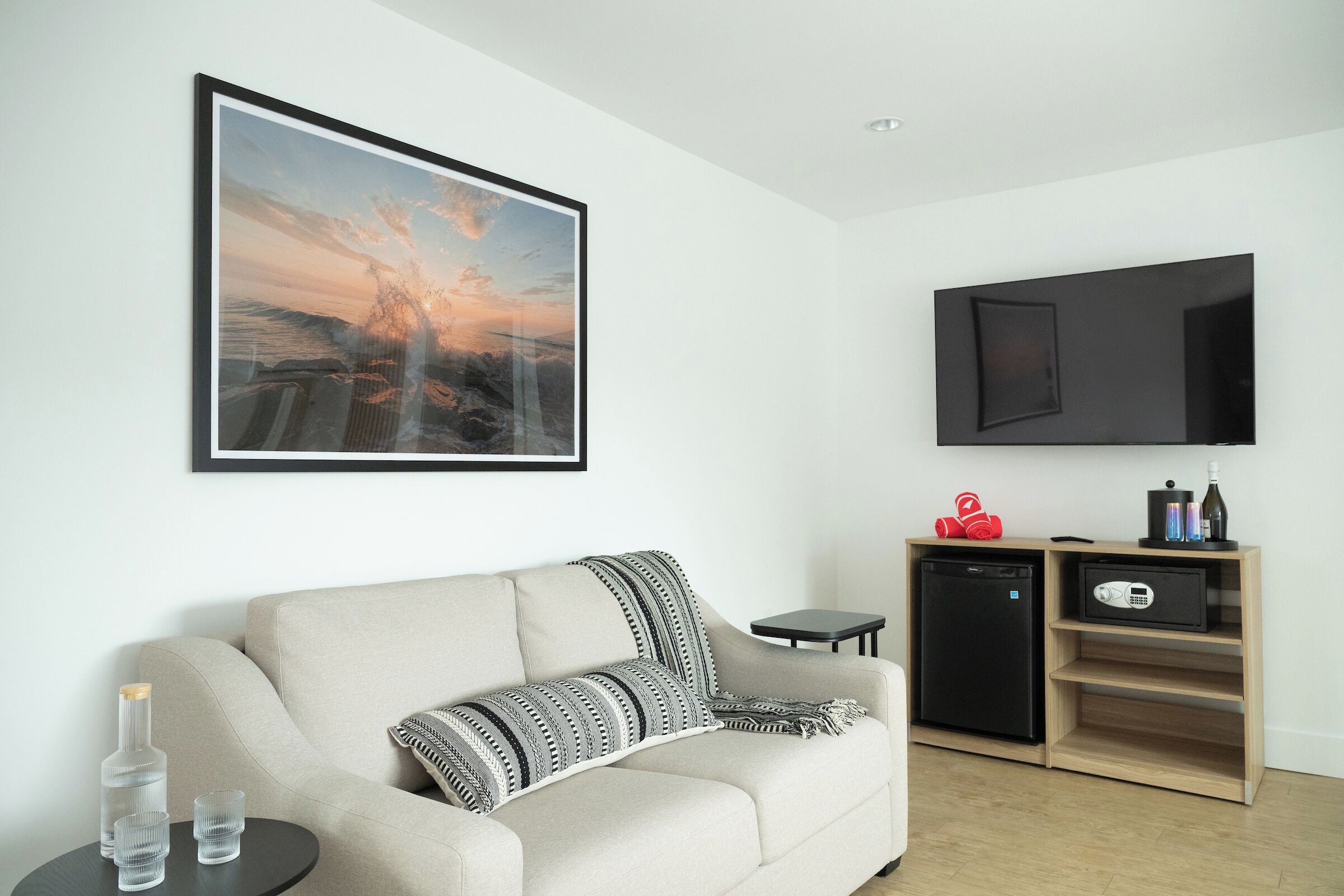 A modern living room with a sofa, a small table with glasses, a framed picture, a TV, and a mini-fridge.
