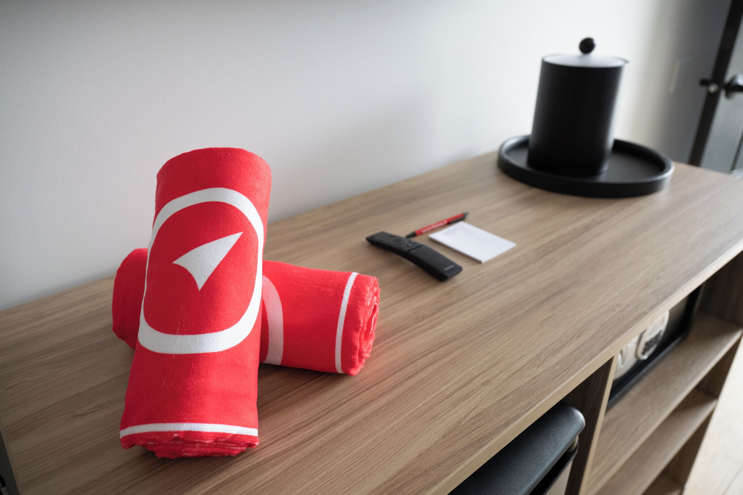 Un bureau en bois sur lequel se trouvent une serviette rouge enroulée, une télécommande noire, un stylo rouge et un récipient noir.