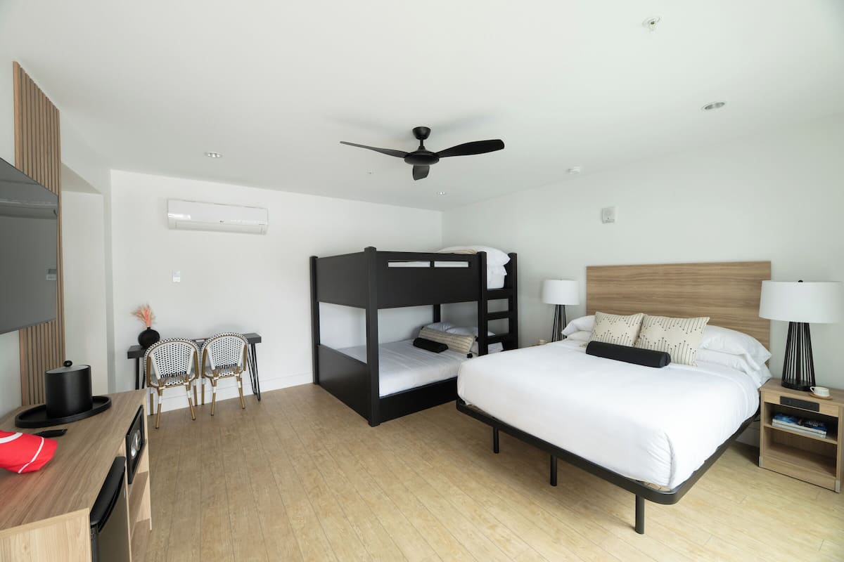 A hotel room with a bunk bed, a desk with a chair, a television, a ceiling fan, and a bedside table with a lamp.
