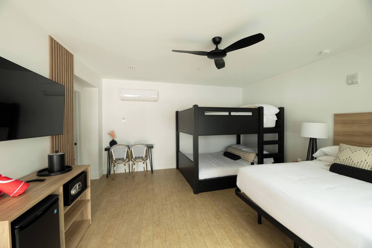A modern hotel room with a bunk bed, a desk with a chair, a television, and a ceiling fan.