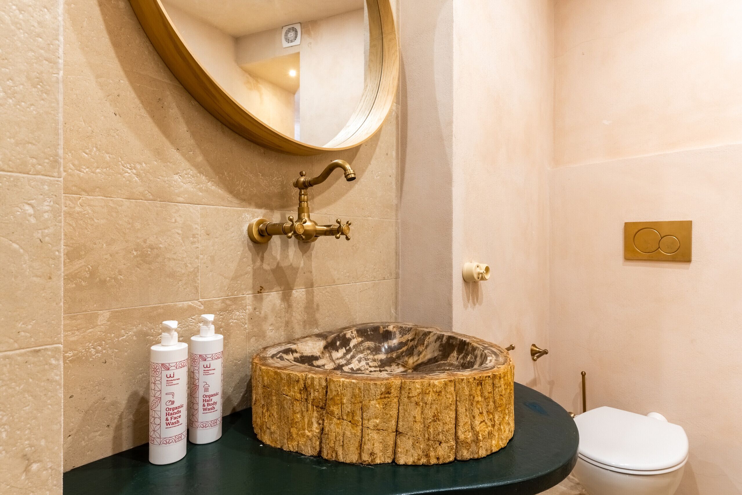 A bathroom with a round wooden basin, two bottles of organic hand and body wash, a gold faucet, and a white toilet.