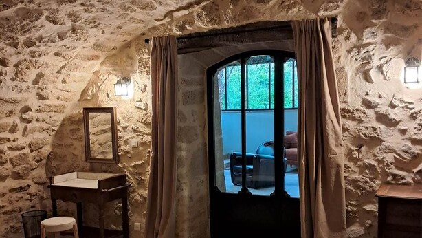Una habitación con paredes de piedra, un ventanal grande, un escritorio de madera, una silla y un taburete pequeño.