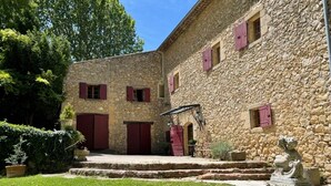 Un edificio de piedra con contraventanas rojas, un pequeño jardín y una estatua.