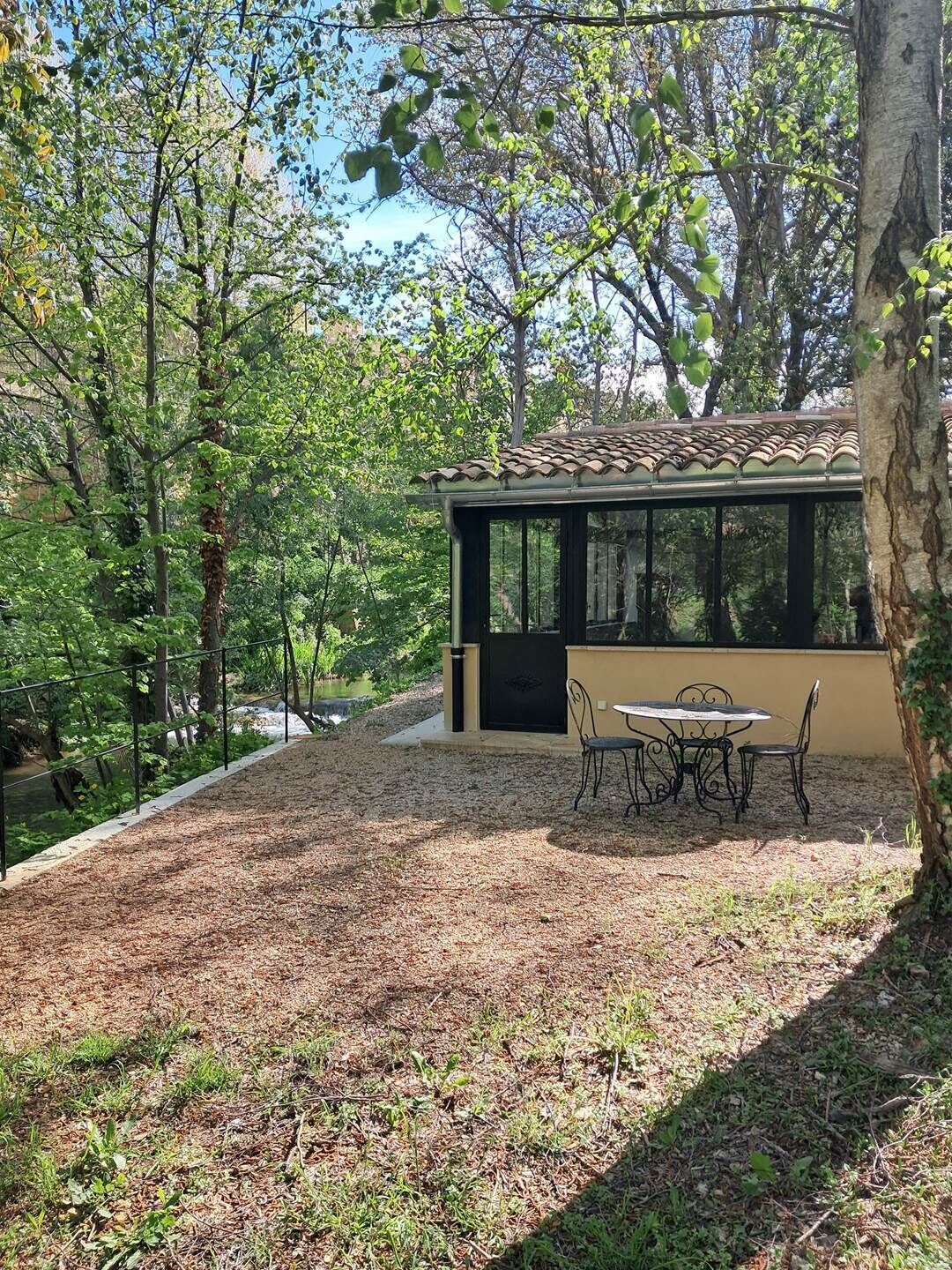 Ein überdachter Außenbereich mit Tisch und Stühlen, umgeben von Bäumen und Blick auf ein Gewässer.