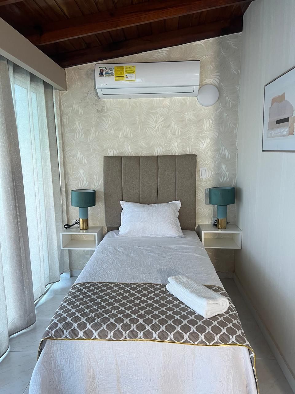 Una cama bien hecha con una colcha estampada, dos lámparas de noche y un equipo de aire acondicionado sobre la cama.