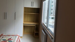 A bedroom with a bed, a window with a view of buildings, and built-in white cabinets with handles. - Ap Beira Mar - Ed Belo Mar - BC (Balneario Camboriu)