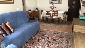 A living room with a blue sofa, a dining table with chairs, a patterned rug, and a painting on the wall. - Ap Beira Mar - Ed Belo Mar - BC (Balneario Camboriu)