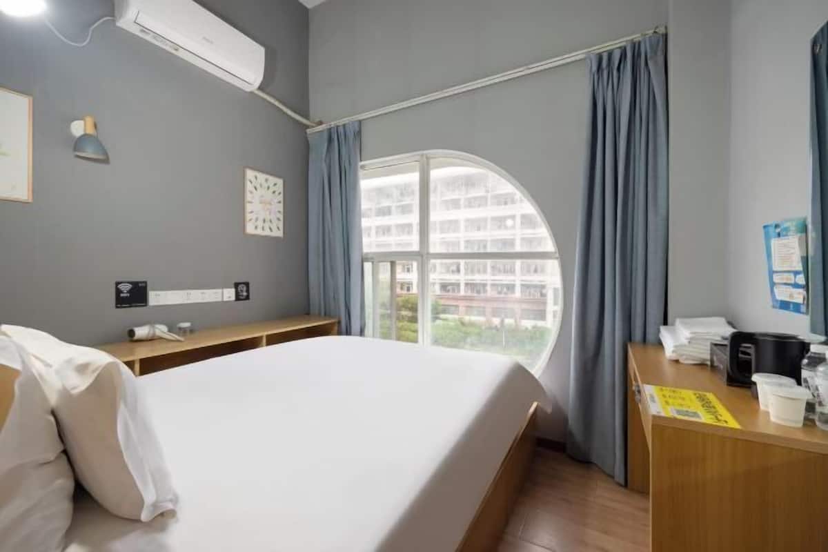A hotel room with a bed, a desk with a telephone, a window with curtains, and a wall-mounted air conditioner.