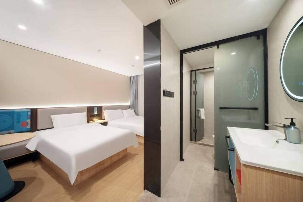 Ein modernes Hotelzimmer mit einem großen Bett, einem Schreibtisch, einem Badezimmer mit Spiegel und Waschbecken sowie einer Glastür zu einem weiteren Raum.