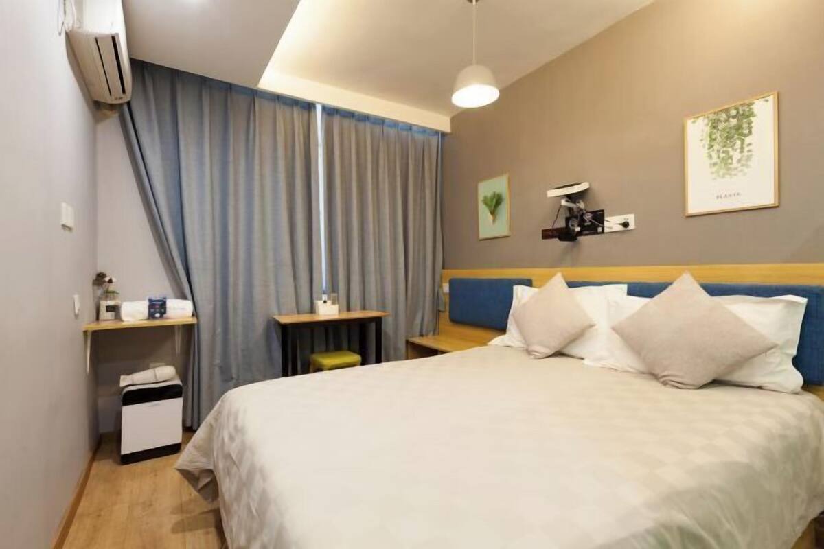 Ein Hotelzimmer mit Bett, Nachttischen, einem Schreibtisch, einer Klimaanlage, einem Fenster mit Vorhängen und Wandbildern.