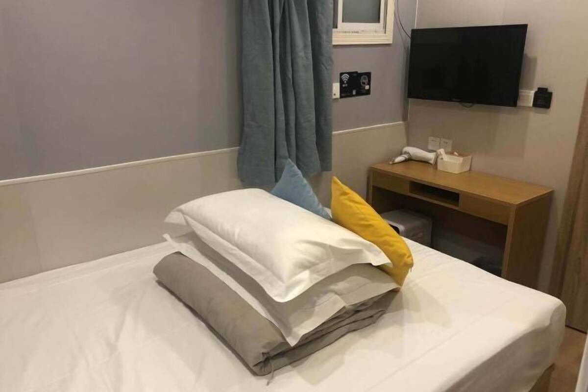 Ein ordentlich bezogenes Bett mit weißer und grauer Bettwäsche, einem gelben und einem grauen Kopfkissen. Ein Nachttisch aus Holz mit einer Lampe und einer Taschentuchbox steht neben dem Bett.