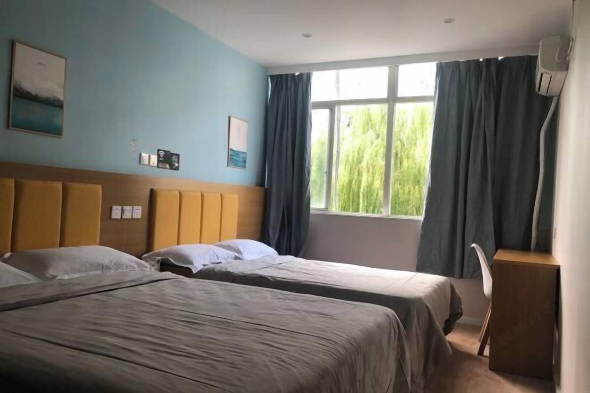 Ein Hotelzimmer mit zwei Betten, einem hölzernen Kopfteil, einem Fenster mit Vorhängen und einem kleinen Schreibtisch mit Stuhl.