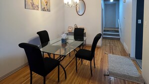 A dining area with a glass table, four black chairs, wooden flooring, a chandelier, and two wall-mounted paintings.