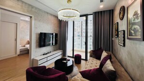 A modern living room with a flat-screen TV, a tufted sofa with purple cushions, a round coffee table, and a chandelier.