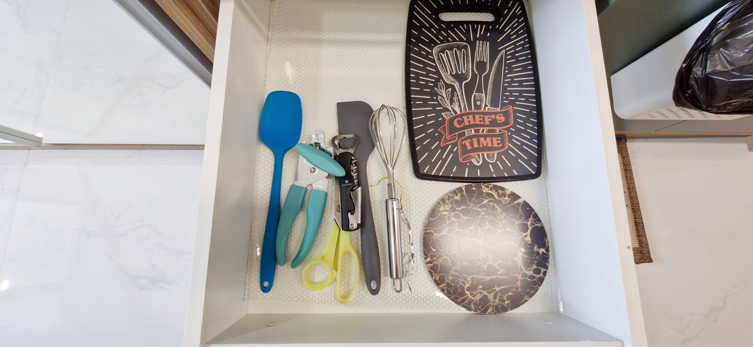 A kitchen drawer with a spatula, knife, whisk, and a chef's time-themed cutting board.