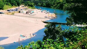 Une plage au bord de la rivière, avec un espace de stationnement, un petit bâtiment et un pont en arrière-plan.
