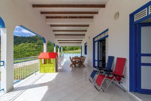 Un balcón techado con una mesa y sillas, una casita de juegos y vistas a un paisaje verde.