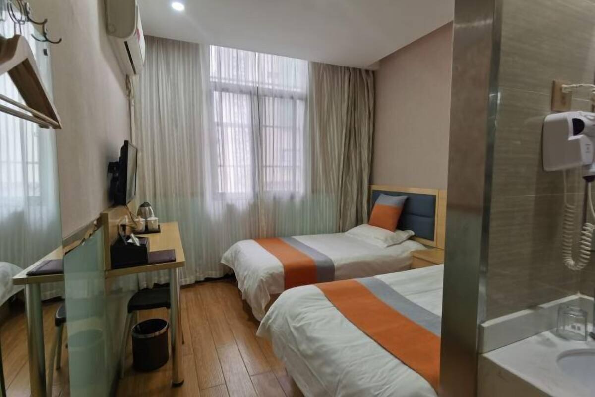 Une chambre d’hôtel avec deux lits, un bureau, une chaise, une poubelle et une fenêtre avec des rideaux.