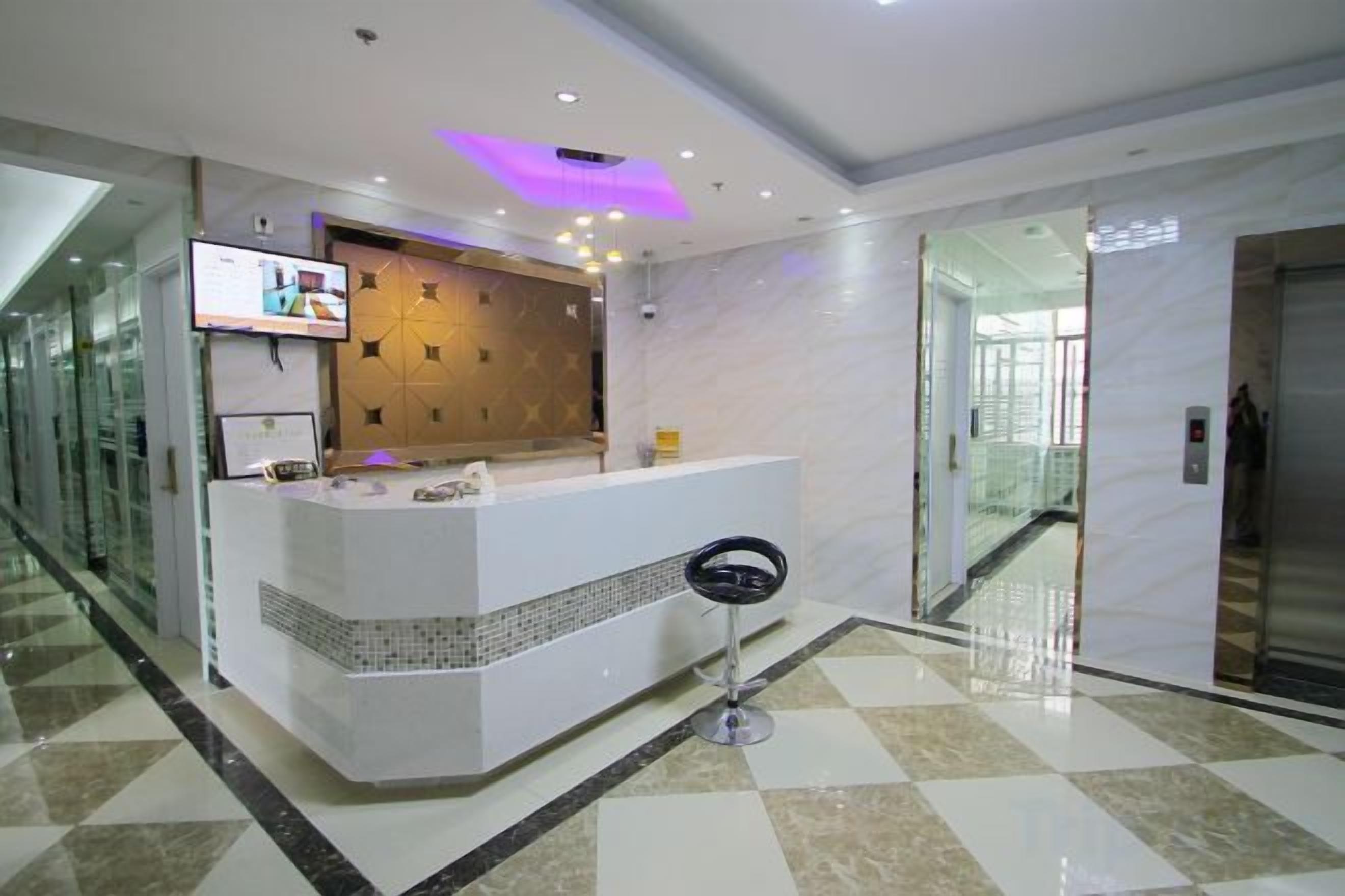 Zona de recepción de un hotel con mostrador, un televisor montado en la pared y puerta de ascensor de vidrio.